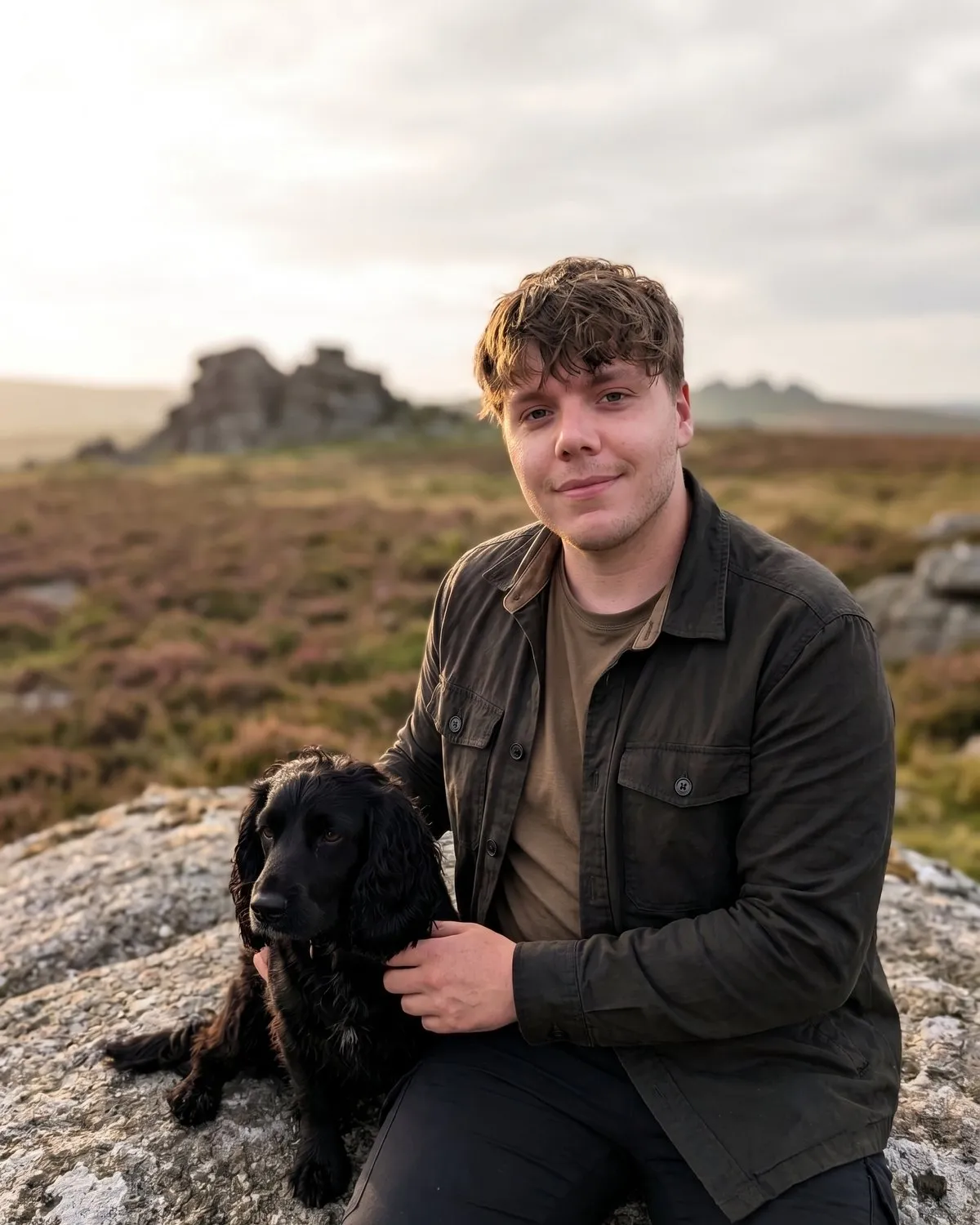 James Willcocks sitting on a granite tor on Dartmoor at golden hour, with his black cocker spaniel Goose beside him